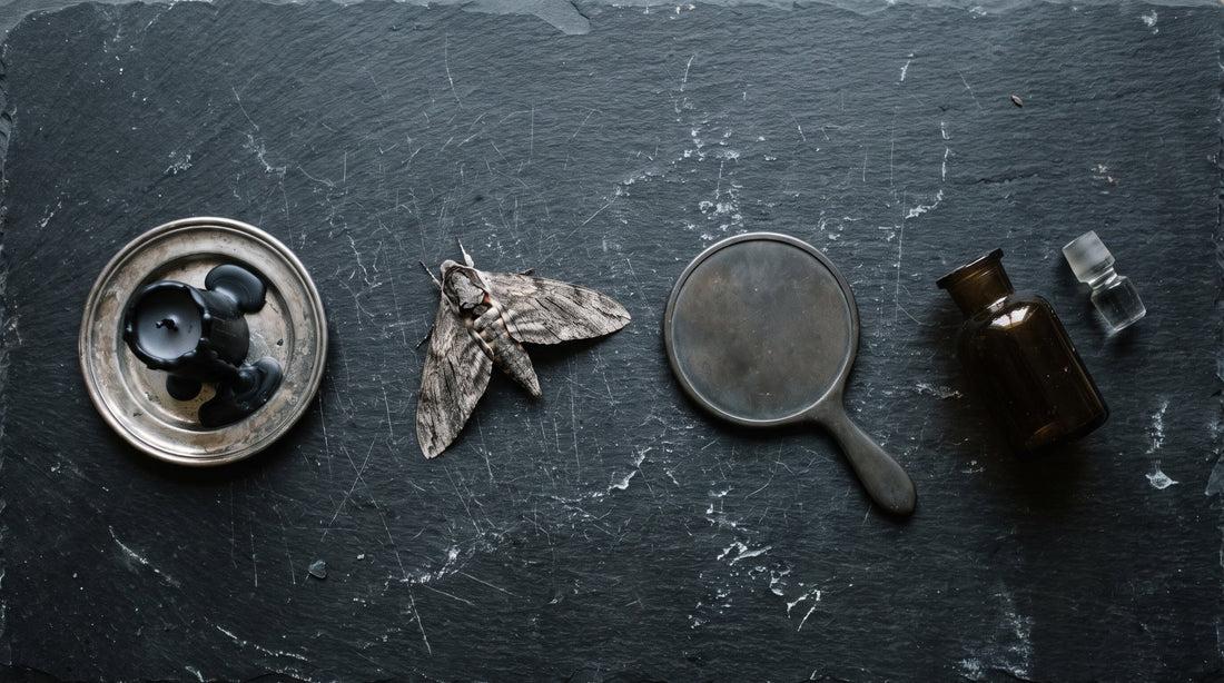 Overhead flat lay of a black candle, a dried moth, a mirror lying face down and a dark apothecary bottle on black slate