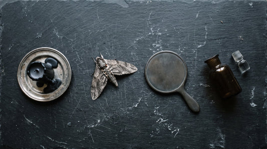 Overhead flat lay of a black candle, a dried moth, a mirror lying face down and a dark apothecary bottle on black slate