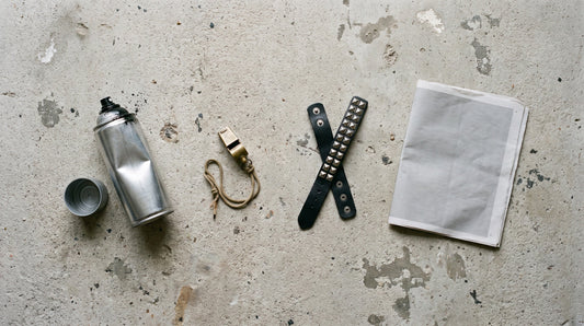 Overhead flat lay of a bare spray can, a brass whistle, a studded leather wristband and a folded blank newspaper on concrete