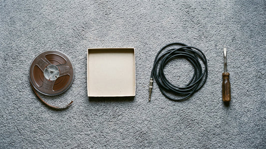 Overhead flat lay of a reel of recording tape, an empty tape box, a coiled jack cable and a screwdriver on studio carpet