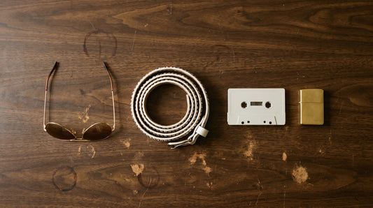 Overhead flat lay of gold aviator sunglasses, a rhinestone studded belt, a blank cassette and a gold lighter on a wood veneer counter
