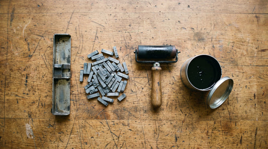 Overhead flat lay of a letterpress composing stick, loose lead type, an inked brayer and a tin of black ink on a printing press bed