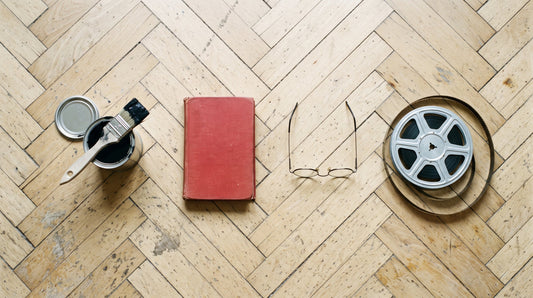 Overhead flat lay of an open tin of black paint with a brush, a red hardback book, round spectacles and a 16mm film reel on a parquet floor