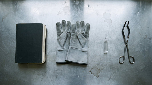 Overhead flat lay of a closed notebook, lead lined gloves, a glass ampoule and steel tongs on a scratched laboratory bench