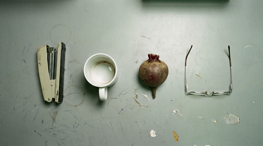 Overhead flat lay of a stapler, an empty mug, a beetroot and a pair of wire rimmed glasses on a grey office desk