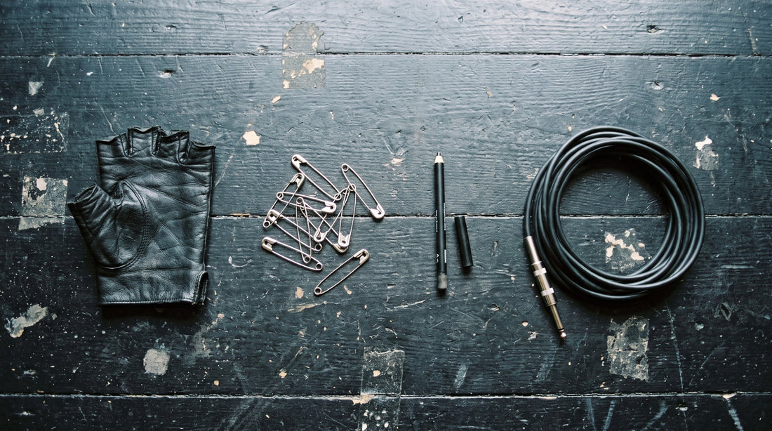 Overhead flat lay of a black leather glove, scattered safety pins, a kohl pencil and a coiled jack cable on black stage boards