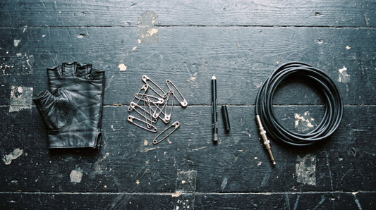 Overhead flat lay of a black leather glove, scattered safety pins, a kohl pencil and a coiled jack cable on black stage boards