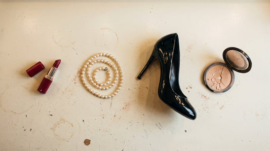 Overhead flat lay of a red lipstick, a strand of pearls, a black patent heel and an open powder compact on a dressing room counter