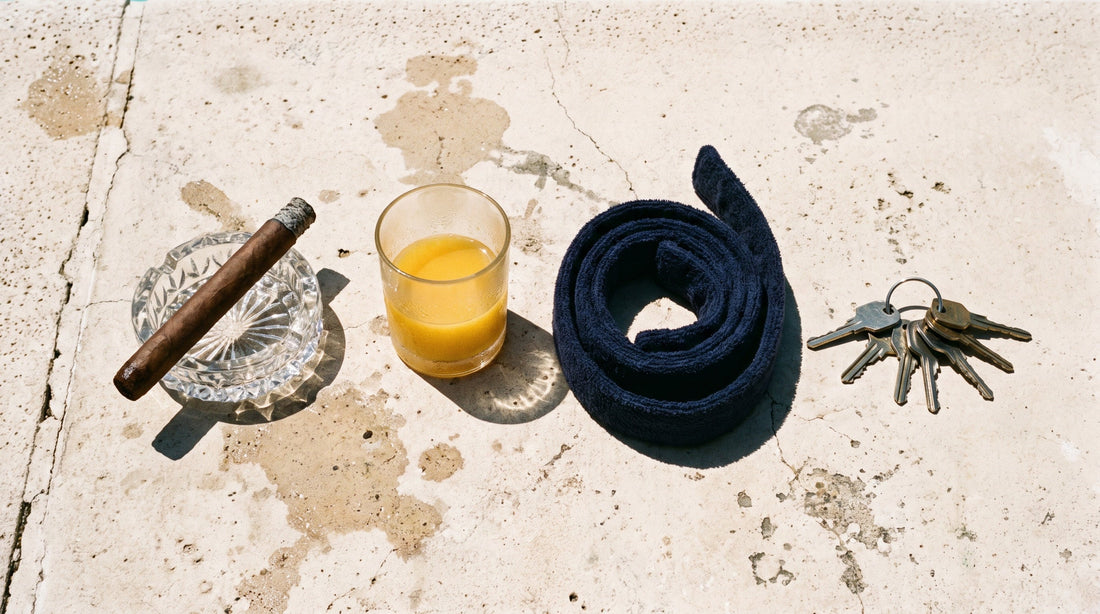 Overhead flat lay of a cigar in a cut glass ashtray, a glass of orange juice, a coiled robe belt and a bunch of car keys on poolside concrete