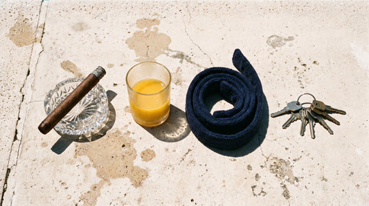 Overhead flat lay of a cigar in a cut glass ashtray, a glass of orange juice, a coiled robe belt and a bunch of car keys on poolside concrete