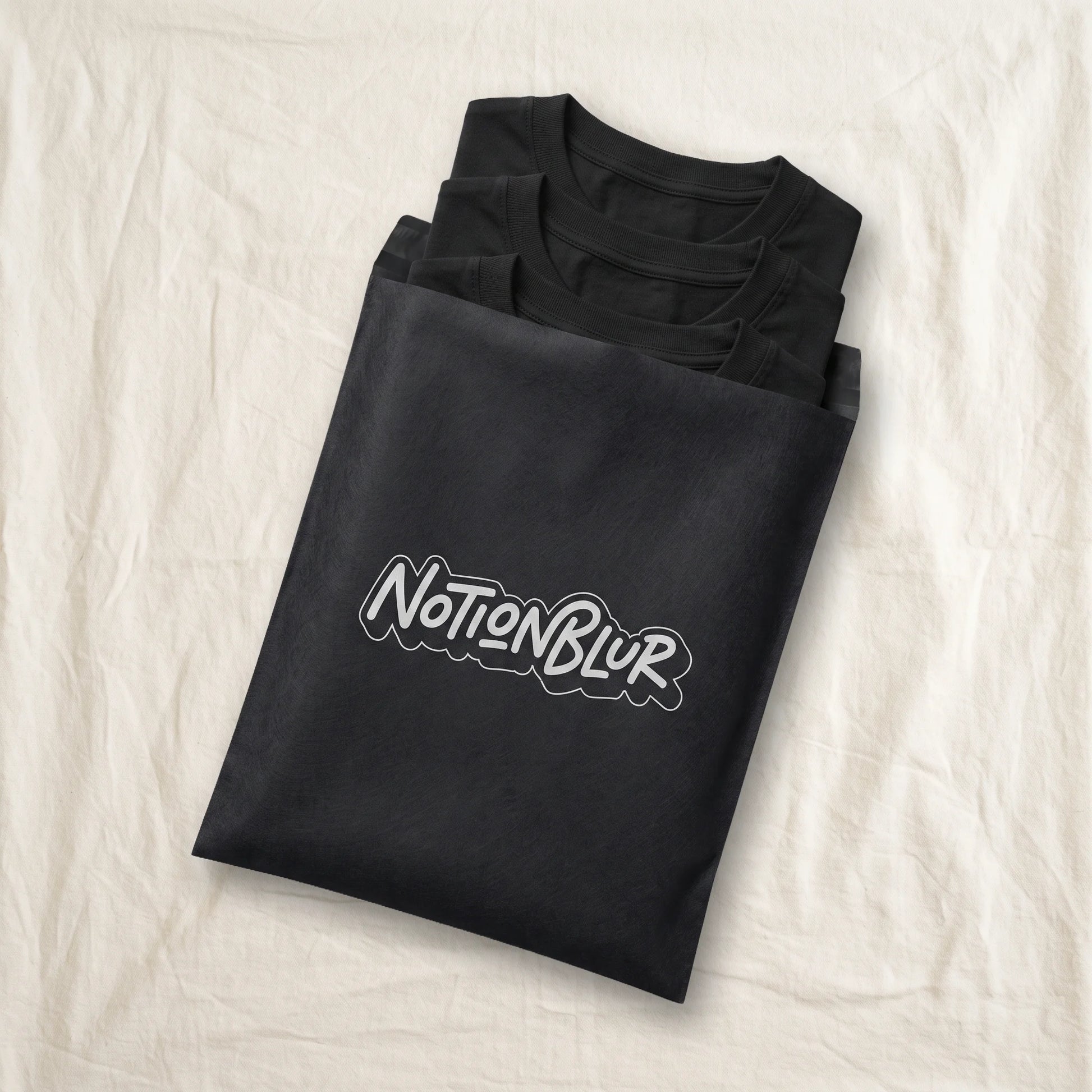 Three folded Notionblur tees in a black mailer on a rumpled cotton bedsheet